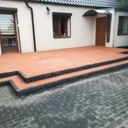 Nowo ułożony taras z czerwonej kostki brukowej z czarnymi obrzeżami, prowadzący do domu z białymi ścianami i ciemną dachówką, otoczony szarą kostką brukową.