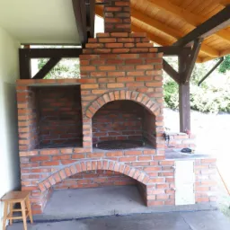 Murowany grill ogrodowy z cegły klinkierowej pod zadaszeniem, z półką, rusztem i małą szafką, widoczny drewniany stołek.