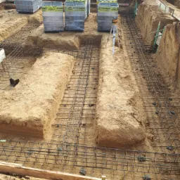 Przygotowanie zbrojenia w wykopie pod fundamenty, widoczne szalunki, stalowe pręty zbrojeniowe, poziomica laserowa na statywie i palety z materiałami budowlanymi w tle.