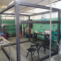 Konstrukcja z profili stalowych w warsztacie, przygotowana do dalszej obróbki, widoczne ślady po spawaniu i otwory montażowe, dwa krzesła wewnątrz konstrukcji, sprzęt warsztatowy w tle.