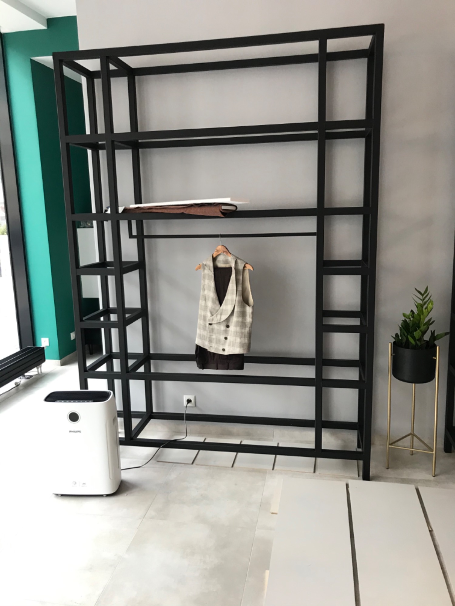 Czarna, metalowa konstrukcja regału z ubraniami na wieszaku, widoczny oczyszczacz powietrza Philips oraz doniczka z rośliną w tle ściana w odcieniach szarości i zieleni.