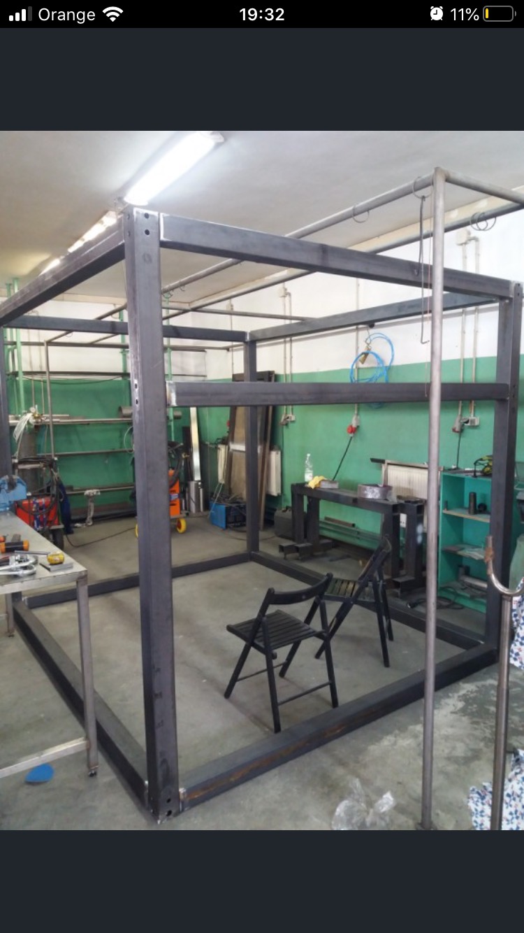 Konstrukcja z profili stalowych w warsztacie, przygotowana do dalszej obróbki, widoczne ślady po spawaniu i otwory montażowe, dwa krzesła wewnątrz konstrukcji, sprzęt warsztatowy w tle.