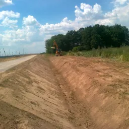 Żółta koparka wykonuje prace ziemne w Oleśnicy, tworząc głęboki rów w piaszczystej glebie. W tle widoczna droga i linia drzew pod błękitnym niebem z chmurami.