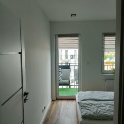 E i J JAROSŁAW KĘDZIORA REMONTY I WYKOŃCZENIA WNĘTRZ - Nowoczesna sypialnia z drewnianą podłogą imitującą deski, białymi ścianami, drzwiami z czarnymi akcentami i widokiem na balkon z sztuczną trawą oraz meblami ogrodowymi przez przesuwne okno...