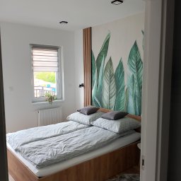 E i J JAROSŁAW KĘDZIORA REMONTY I WYKOŃCZENIA WNĘTRZ - Sypialnia z łóżkiem małżeńskim na drewnianej ramie, szare poduszki i pościel w delikatny wzór, ściana z dekoracyjną tapetą w motywie tropikalnych liści, drewniane lamele, okno z roletami dzień-noc...