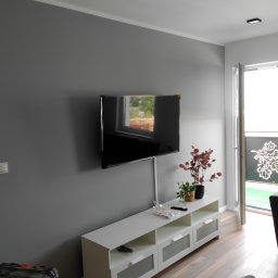 E i J JAROSŁAW KĘDZIORA REMONTY I WYKOŃCZENIA WNĘTRZ - Telewizor wiszący na szarej ścianie nad białą szafką RTV z roślinami doniczkowymi, widoczny kabel zasilający, podłoga z paneli drewnopodobnych, czarna skrzynka narzędziowa w rogu.