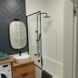 E i J JAROSŁAW KĘDZIORA REMONTY I WYKOŃCZENIA WNĘTRZ - Nowoczesna łazienka z umywalką nablatową na drewnianej szafce, szarą ścianą z dekoracyjnymi płytkami chevron, kabiną prysznicową z czarnymi ramami i białą wanną, widoczna pralka z boku.