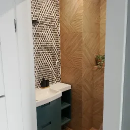 Nowoczesna łazienka z umywalką na szafce w kolorze butelkowej zieleni, mozaiką na ścianie i drewnopodobnymi płytkami ułożonymi w geometryczny wzór.