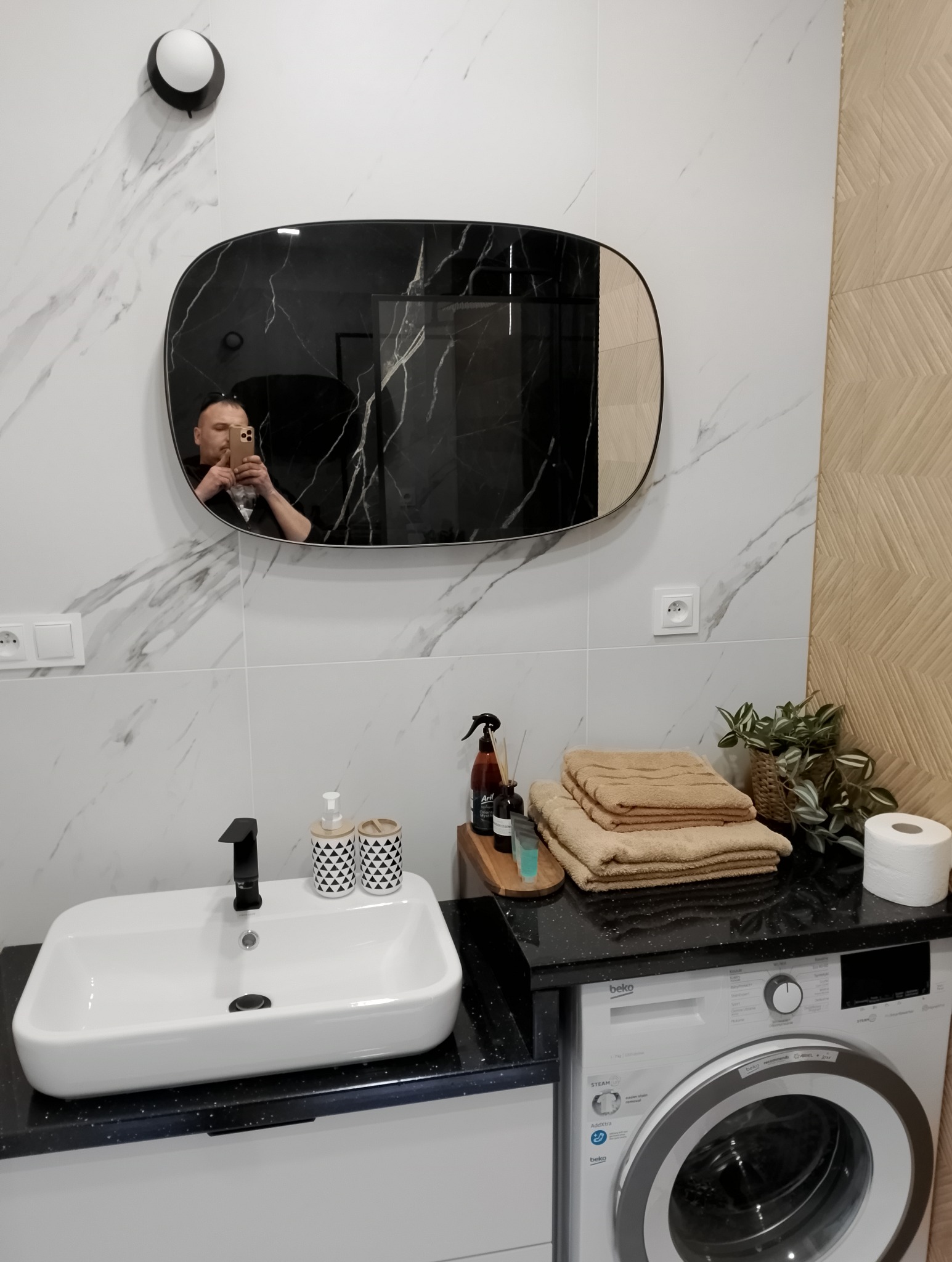 Nowoczesna łazienka z owalnym lustrem z czarnym marmurkowym wzorem, białą umywalką nablatową i pralką Beko w zabudowie, sfotografowana z odbiciem fotografa w lustrze.