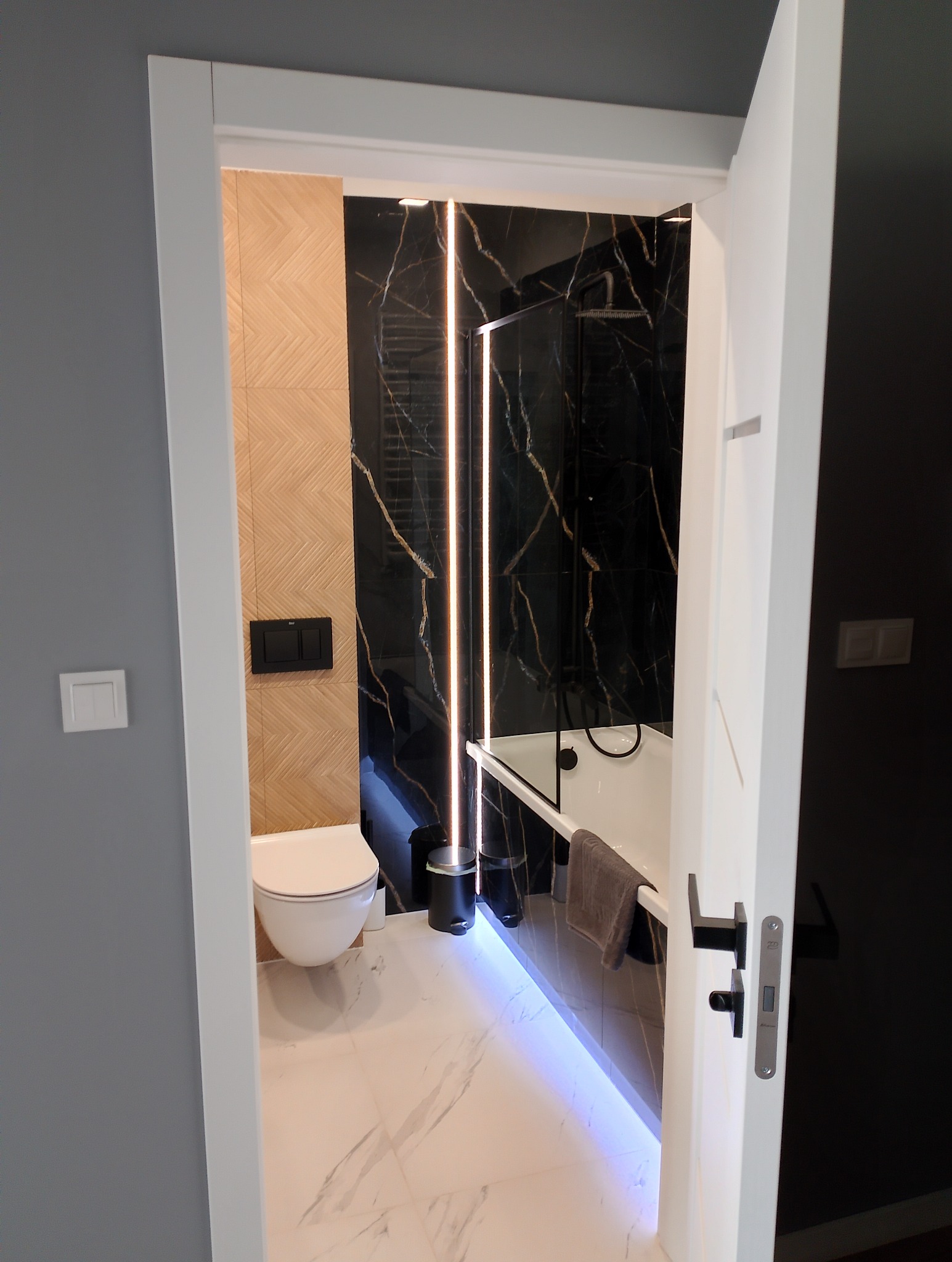 Nowoczesna łazienka z czarnymi marmurowymi płytkami, oświetleniem LED i wiszącą toaletą, widoczna zza otwartych białych drzwi.