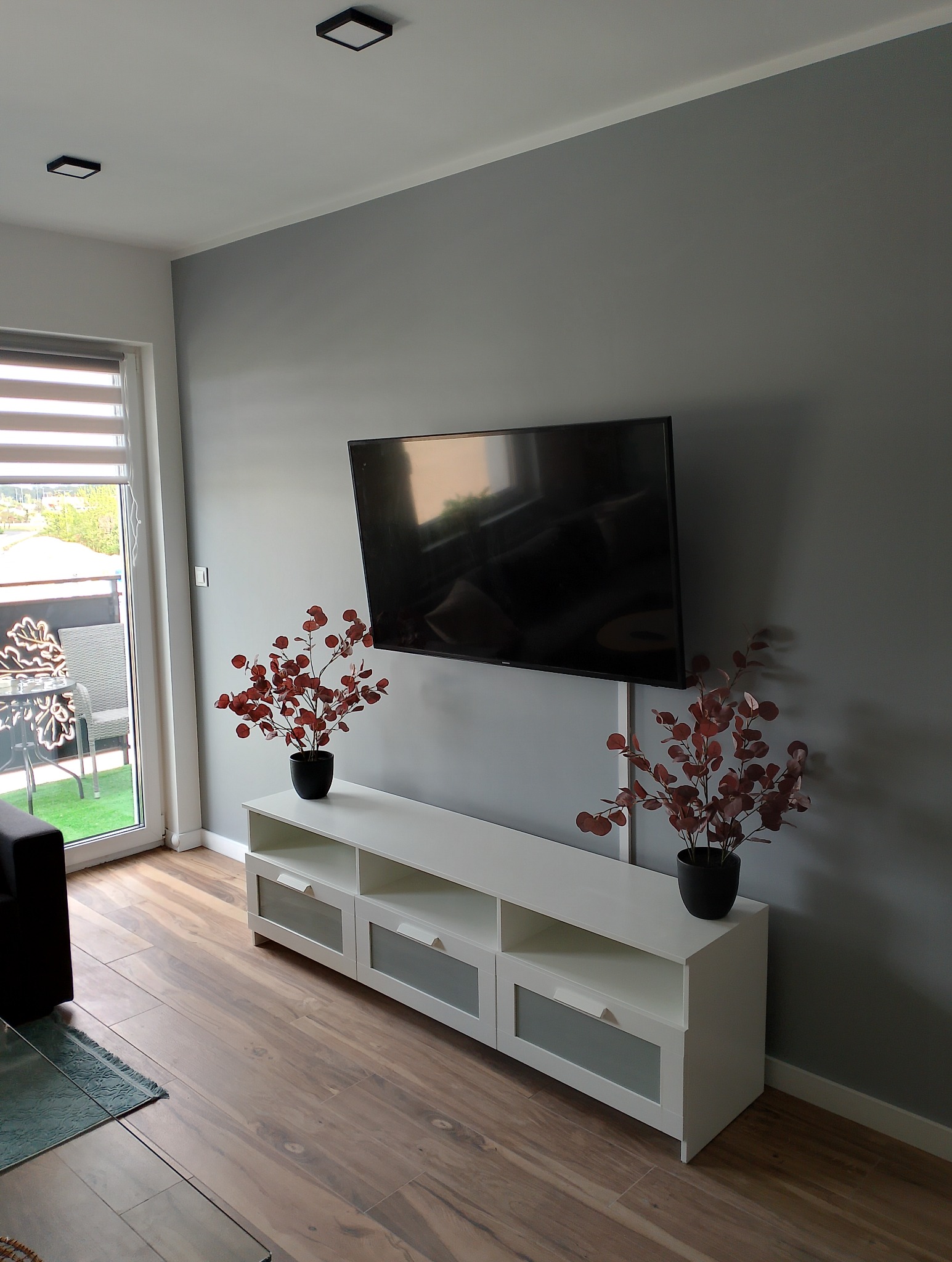 Telewizor zawieszony na szarej ścianie nad białą szafką RTV z trzema szufladami, ozdobiony dwoma bukietami z czerwonymi liśćmi w czarnych wazonach. Na podłodze panele imitujące drewno. Widoczny...