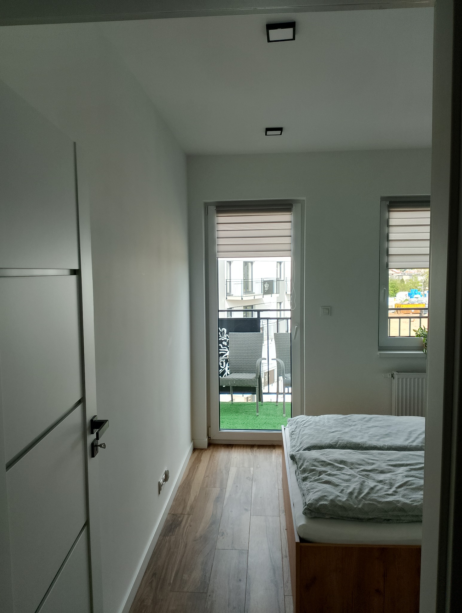 Nowoczesna sypialnia z drewnianą podłogą imitującą deski, białymi ścianami, drzwiami z czarnymi akcentami i widokiem na balkon z sztuczną trawą oraz meblami ogrodowymi przez przesuwne okno...