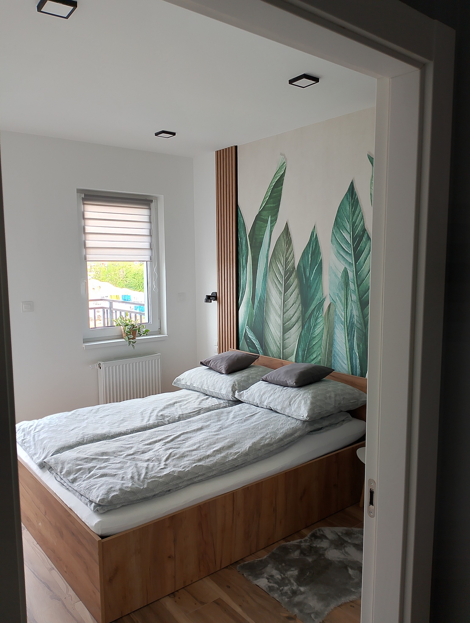 Sypialnia z łóżkiem małżeńskim na drewnianej ramie, szare poduszki i pościel w delikatny wzór, ściana z dekoracyjną tapetą w motywie tropikalnych liści, drewniane lamele, okno z roletami dzień-noc...