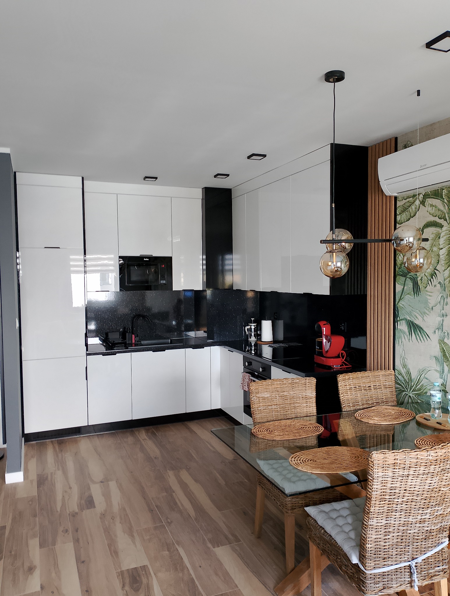 Nowoczesna kuchnia z białymi szafkami, czarnym blatem i okładziną, drewnianą podłogą oraz stołem z szklanym blatem i wiklinowymi krzesłami, oświetlona lampą z kulistymi kloszami
