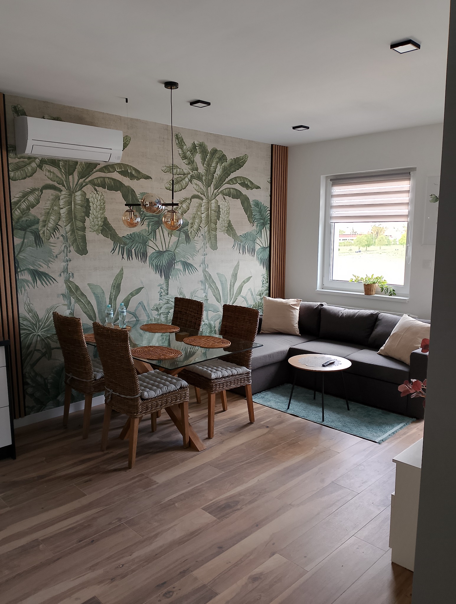 Jasne wnętrze jadalni z motywem tropikalnym na tapecie, szklany stół z rattanowymi krzesłami, drewnopodobna podłoga, szary narożnik z poduszkami i widoczny klimatyzator na ścianie.