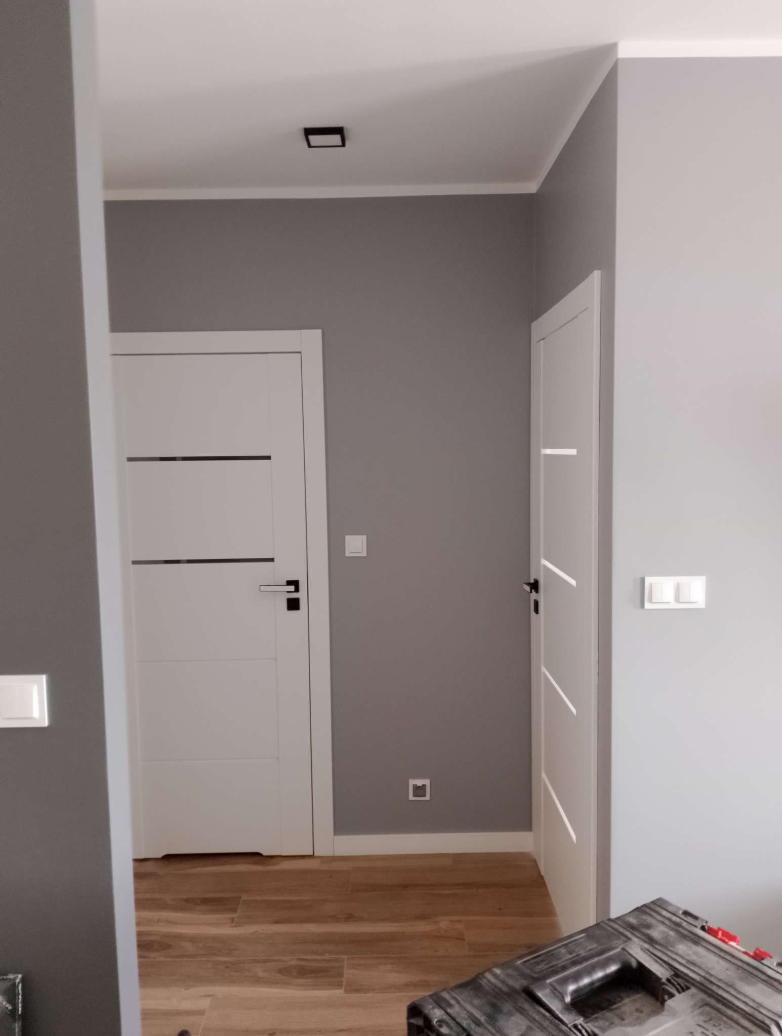 Dwa białe, nowoczesne drzwi z czarnymi klamkami w szarym wnętrzu, widoczne fragmenty podłogi z paneli i skrzynki narzędziowej.