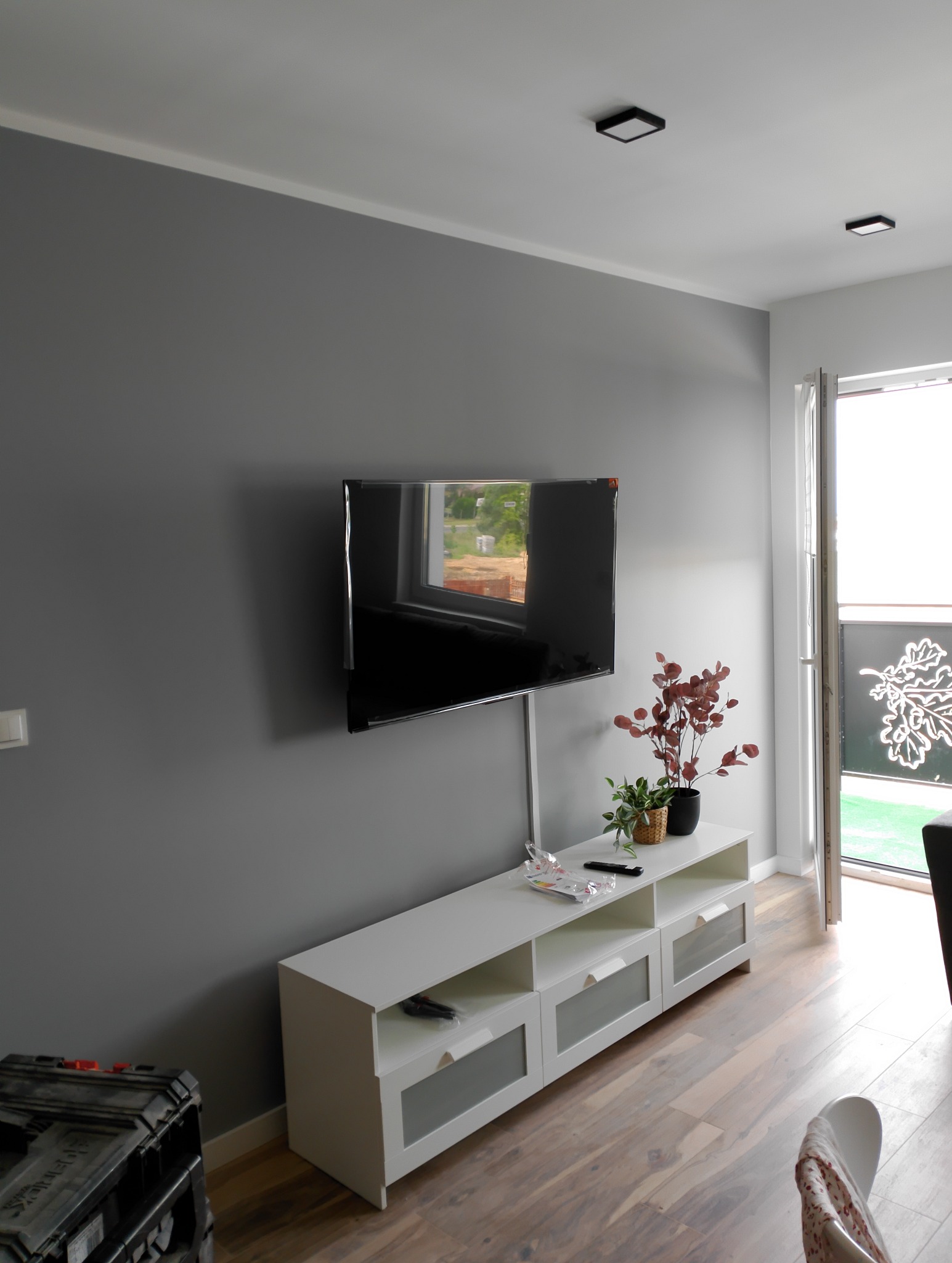 Telewizor wiszący na szarej ścianie nad białą szafką RTV z roślinami doniczkowymi, widoczny kabel zasilający, podłoga z paneli drewnopodobnych, czarna skrzynka narzędziowa w rogu.