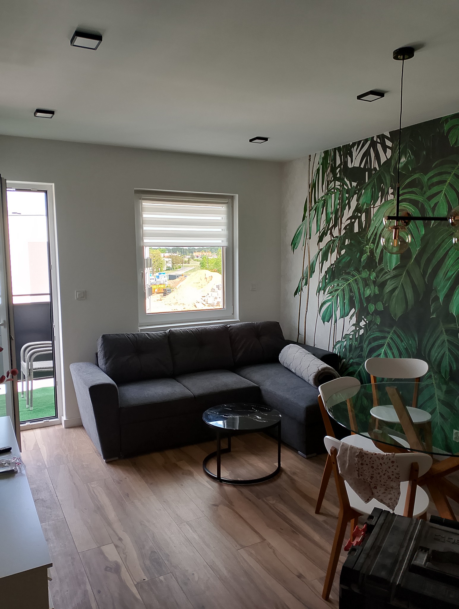 Nowoczesny salon z szarą sofą narożną, okrągłym stolikiem kawowym z czarnym blatem imitującym marmur, drewnianą podłogą i fototapetą z motywem tropikalnych liści na ścianie. W tle widoczne okno...