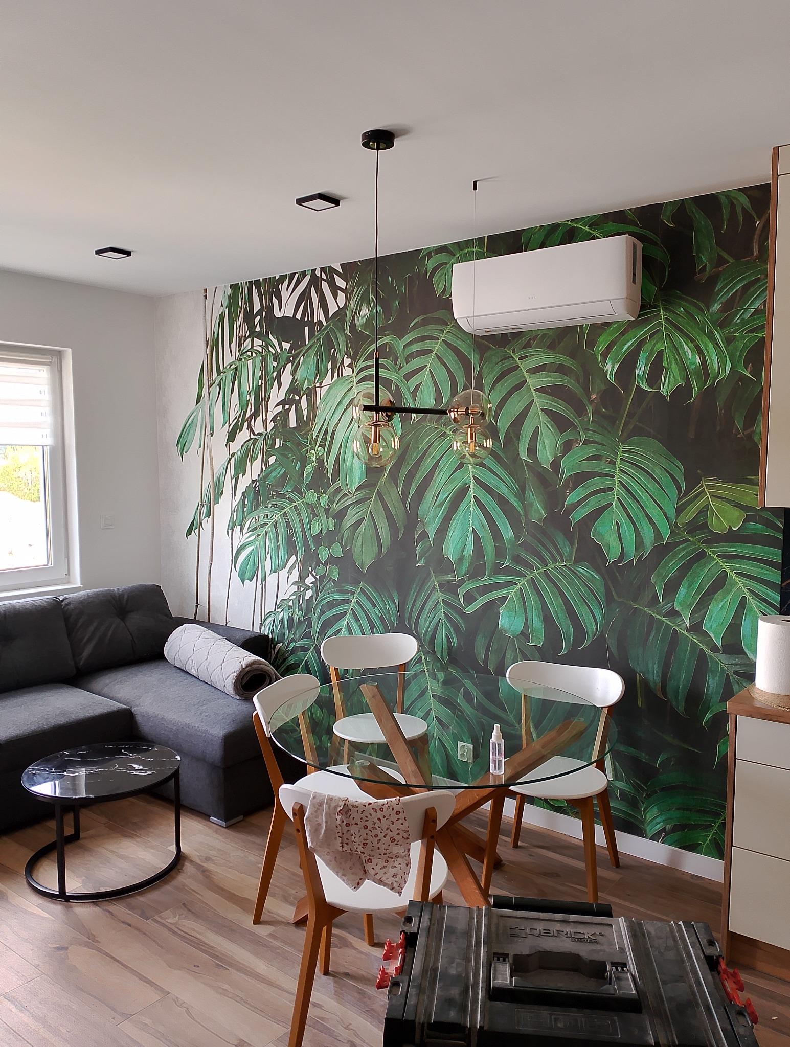 Jasne wnętrze salonu z narożnikiem, okrągłym stołem z szklanym blatem i fototapetą z motywem tropikalnych liści monstery, z widocznym klimatyzatorem i skrzynką narzędziową na podłodze.
