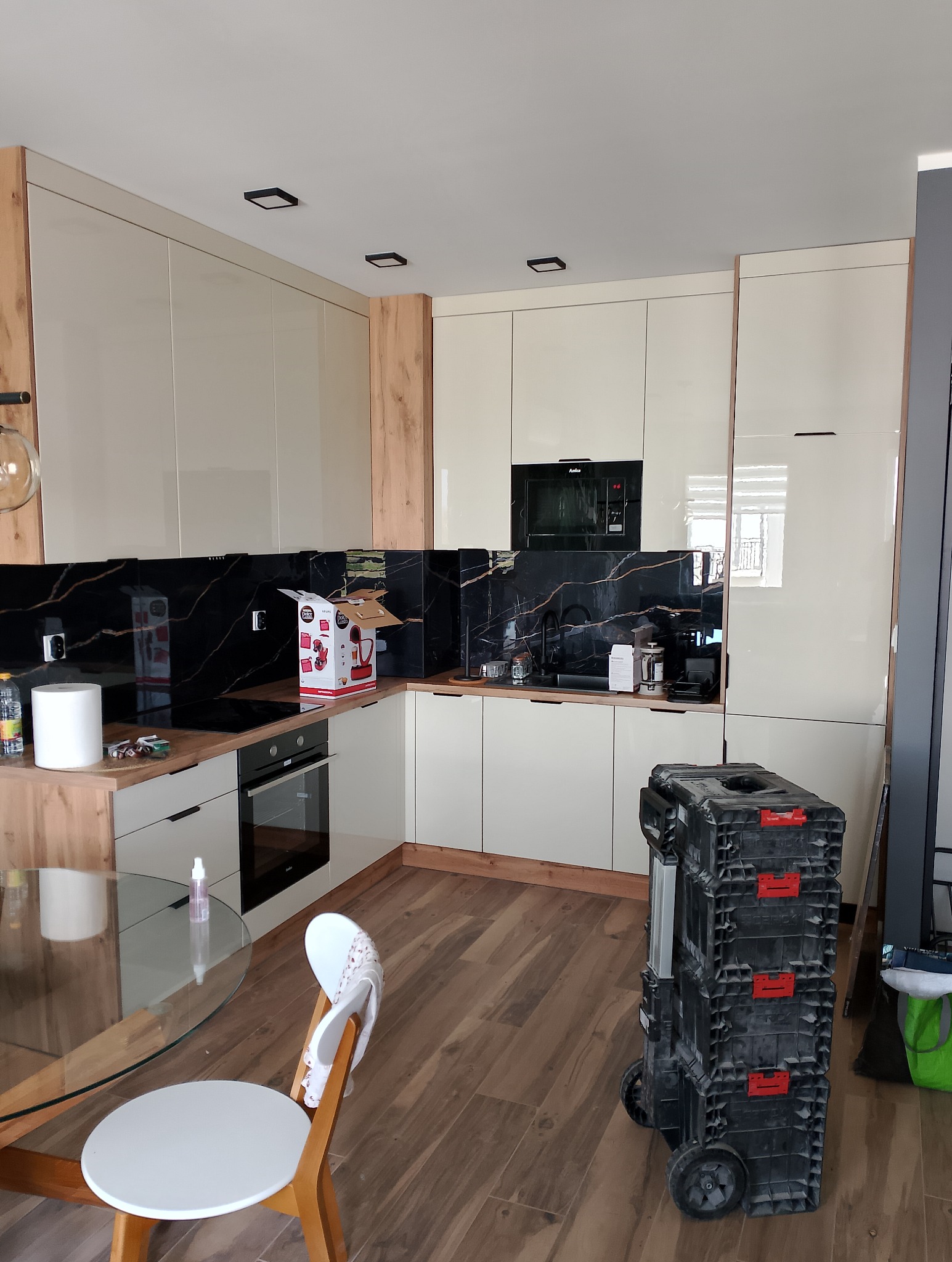 Jasna kuchnia z czarnym marmurowym backsplashiem i drewnianymi akcentami, widoczny zestaw narzędzi na kółkach, szklany stół z krzesłem i drobne akcesoria kuchenne.