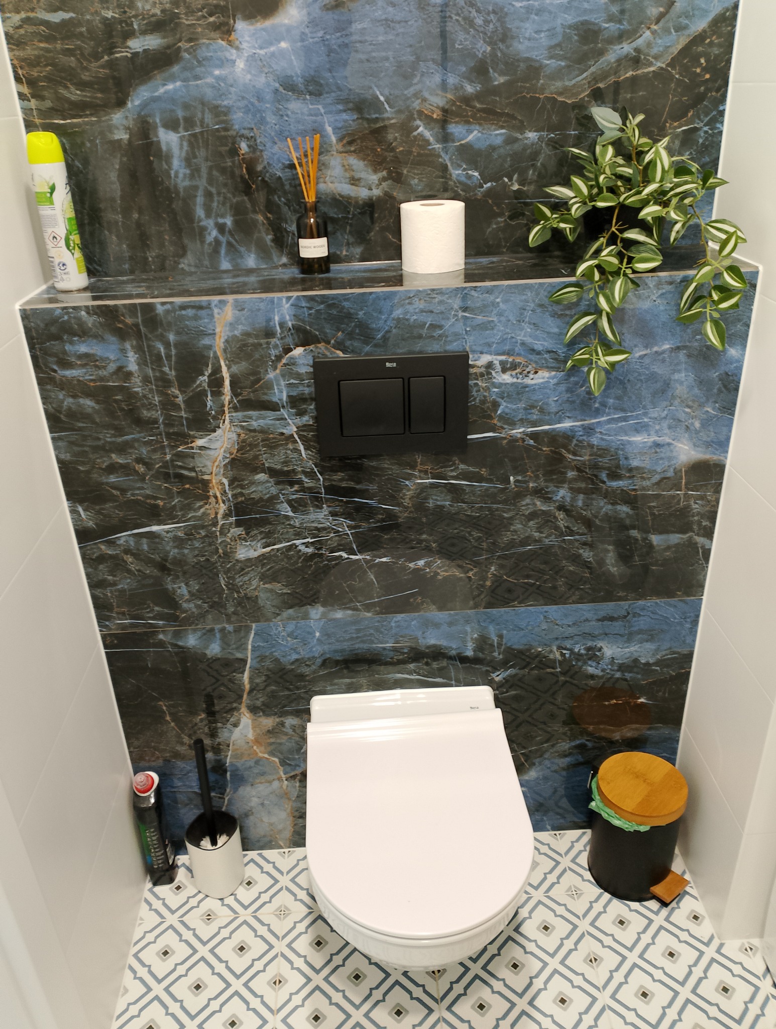 Nowoczesna toaleta podwieszana z czarnym przyciskiem spłukującym na tle płytek imitujących marmur, uzupełniona o roślinę doniczkową, odświeżacz powietrza i elegancki kosz na śmieci z drewnianą...