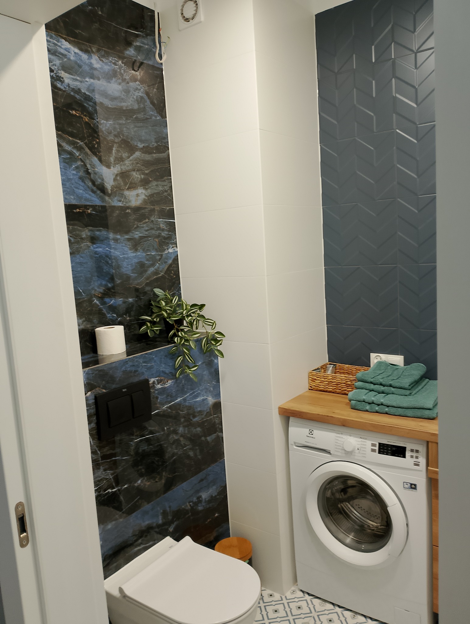 Nowoczesna łazienka z marmurową ścianą, toaletą podwieszaną, pralką Electrolux pod drewnianym blatem z ręcznikami i dekoracyjnymi płytkami w jodełkę.