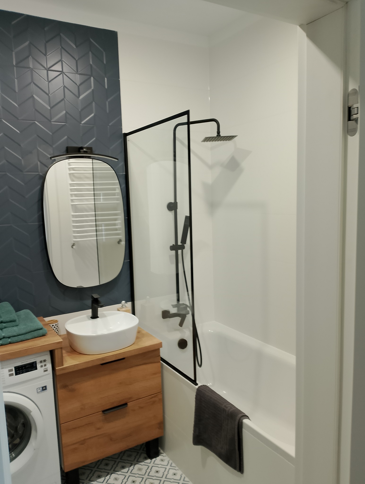 Nowoczesna łazienka z umywalką nablatową na drewnianej szafce, szarą ścianą z dekoracyjnymi płytkami chevron, kabiną prysznicową z czarnymi ramami i białą wanną, widoczna pralka z boku.