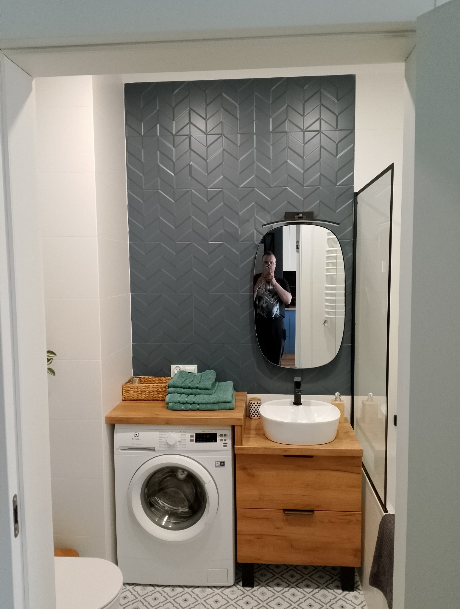 Nowoczesna łazienka z szarą ścianą z płytek ułożonych w jodełkę, drewnianym blatem z umywalką nablatową, pralką Electrolux i lustrem owalnym, odbicie fotografa.