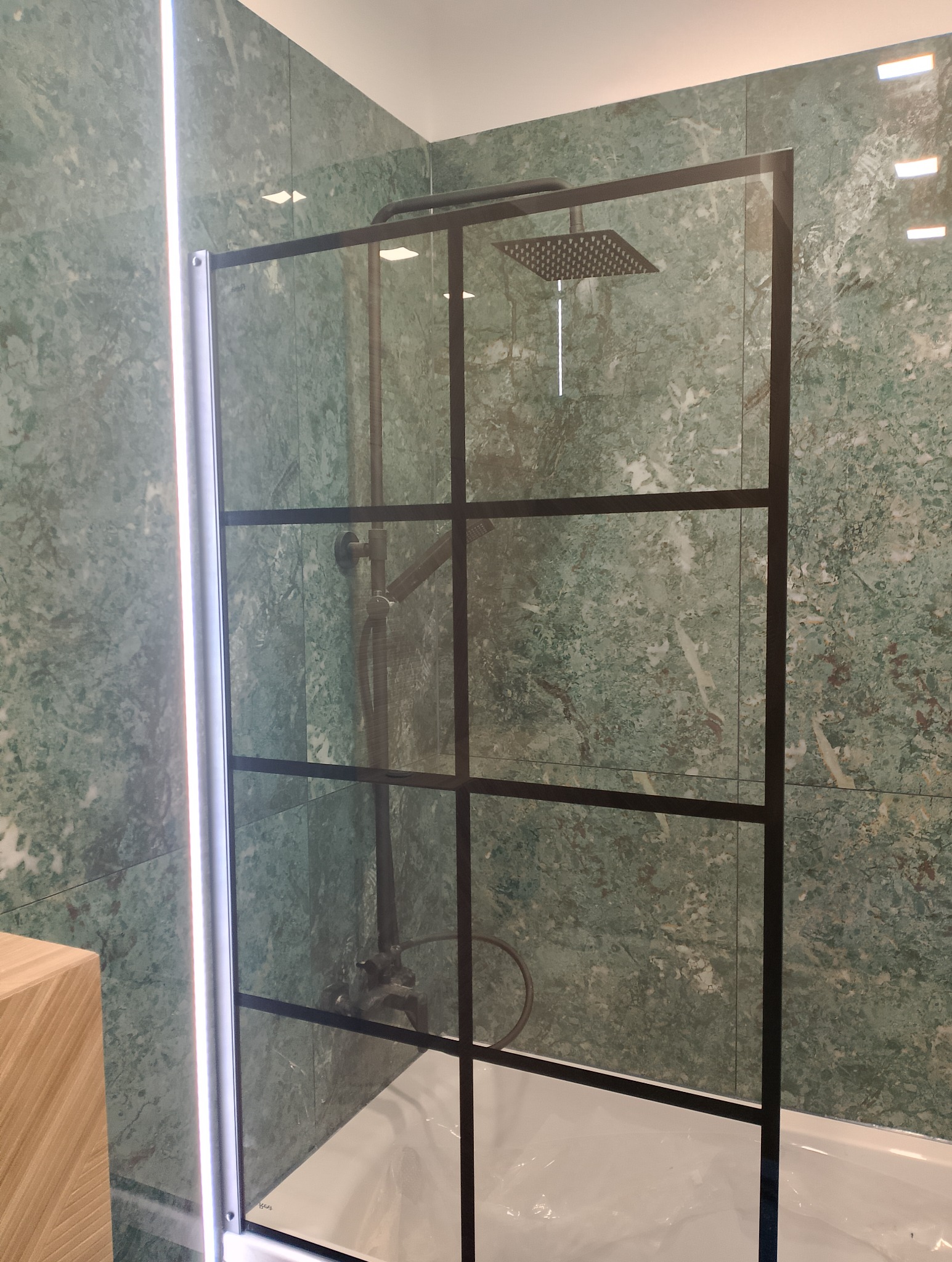 Kabina prysznicowa typu walk-in z czarnymi szprosami na tle ściany wyłożonej zielonym marmurem, widoczny zestaw prysznicowy z deszczownicą