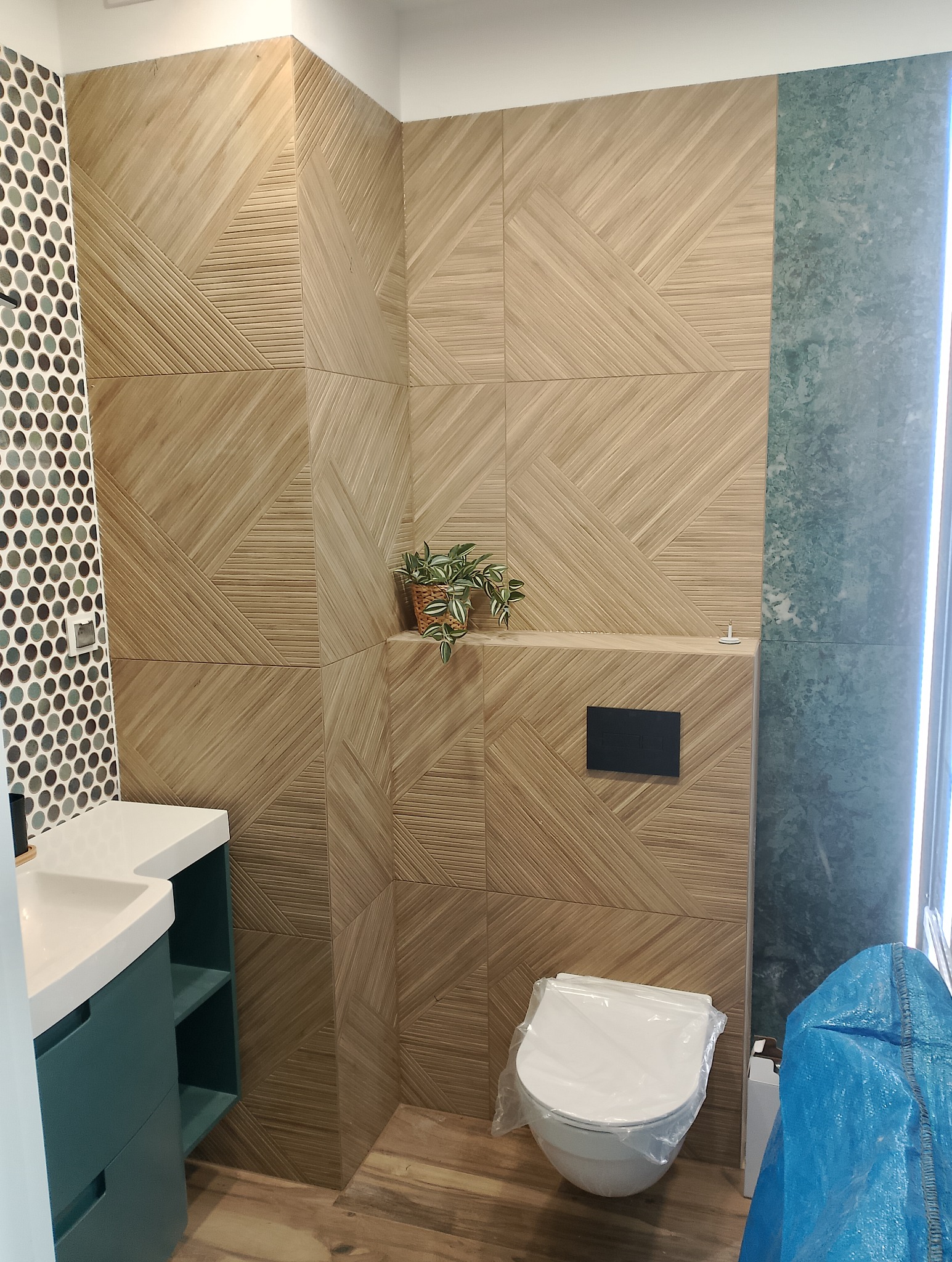 Nowoczesna łazienka z drewnopodobnymi płytkami ułożonymi w geometryczny wzór, okrągłe mozaiki przy umywalce i wisząca toaleta zabezpieczona folią ochronną.