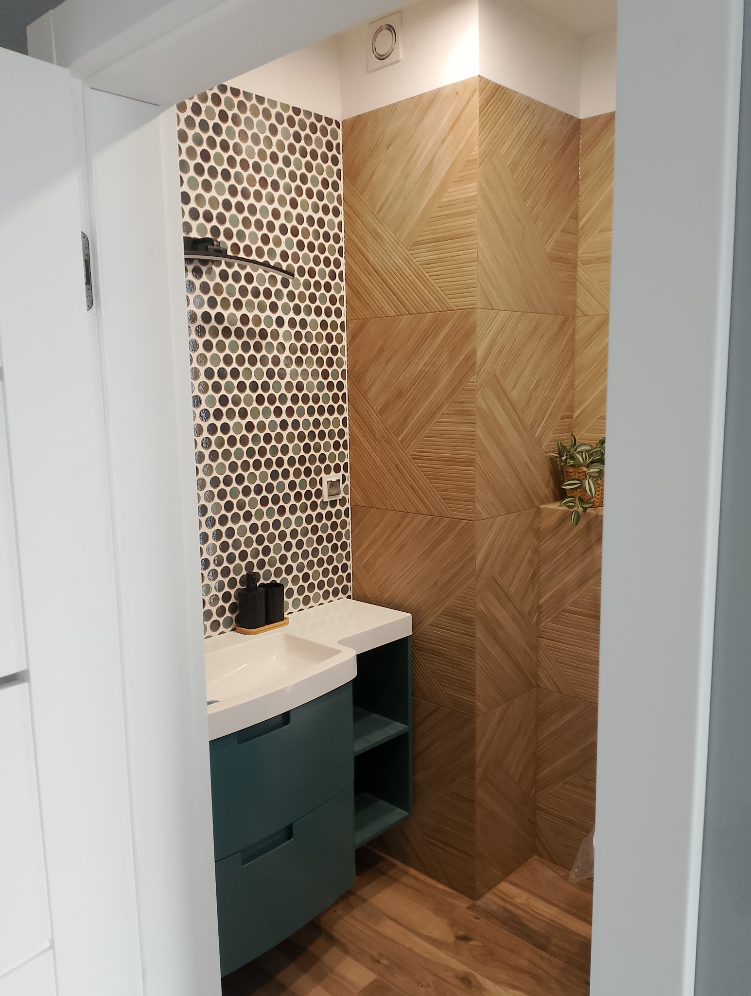 Nowoczesna łazienka z umywalką na szafce w kolorze butelkowej zieleni, mozaiką na ścianie i drewnopodobnymi płytkami ułożonymi w geometryczny wzór.
