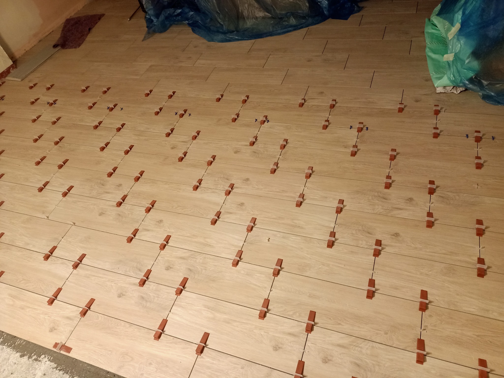 Podłoga z paneli imitujących drewno w trakcie układania, z widocznymi krzyżykami dystansowymi i klinami poziomującymi, zabezpieczona folią malarską w rogu pomieszczenia.