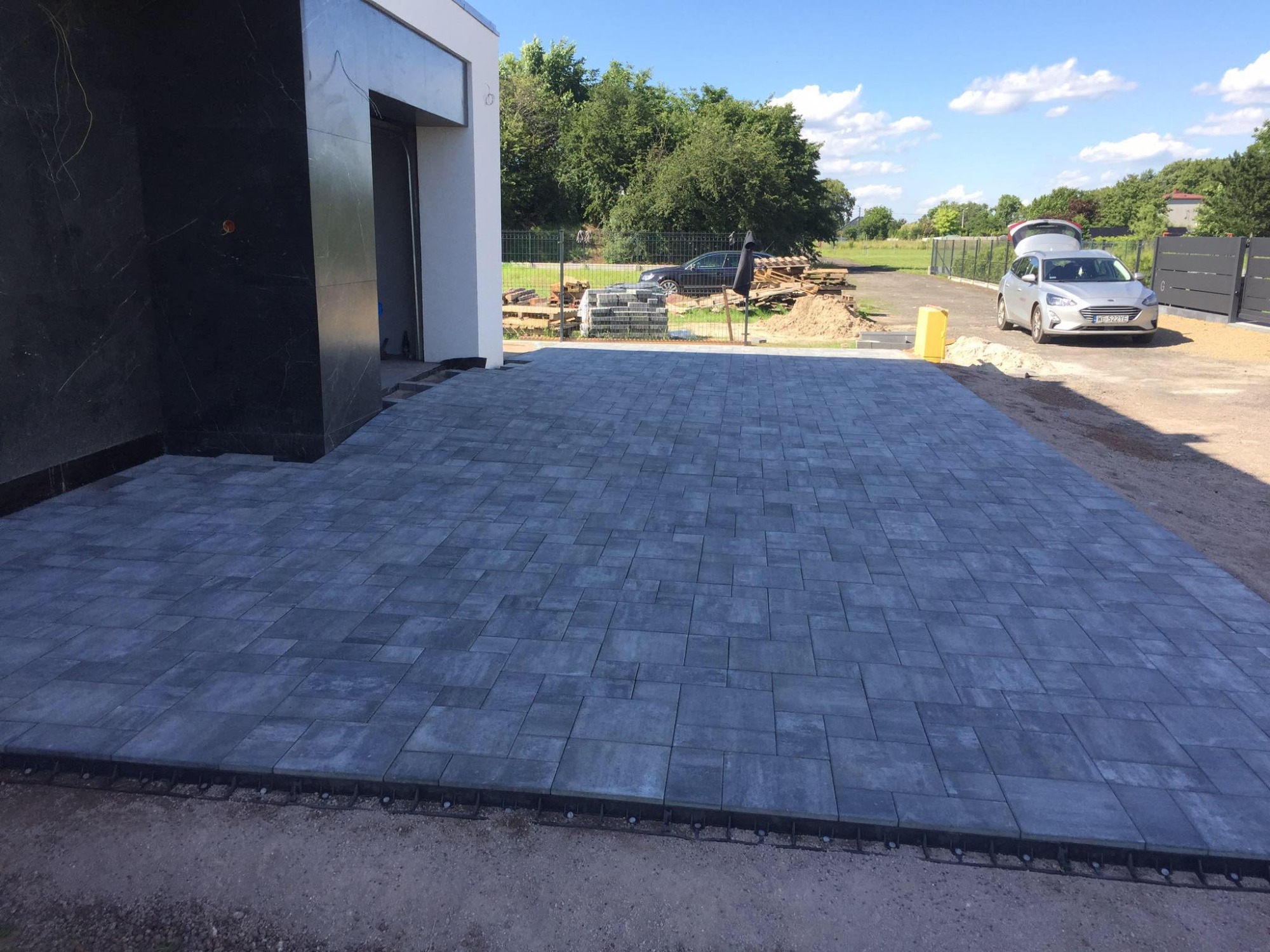 Ułożona, szara kostka brukowa na podjeździe przed nowoczesnym domem z czarną elewacją, widoczne palety z kostką i samochód osobowy w tle.