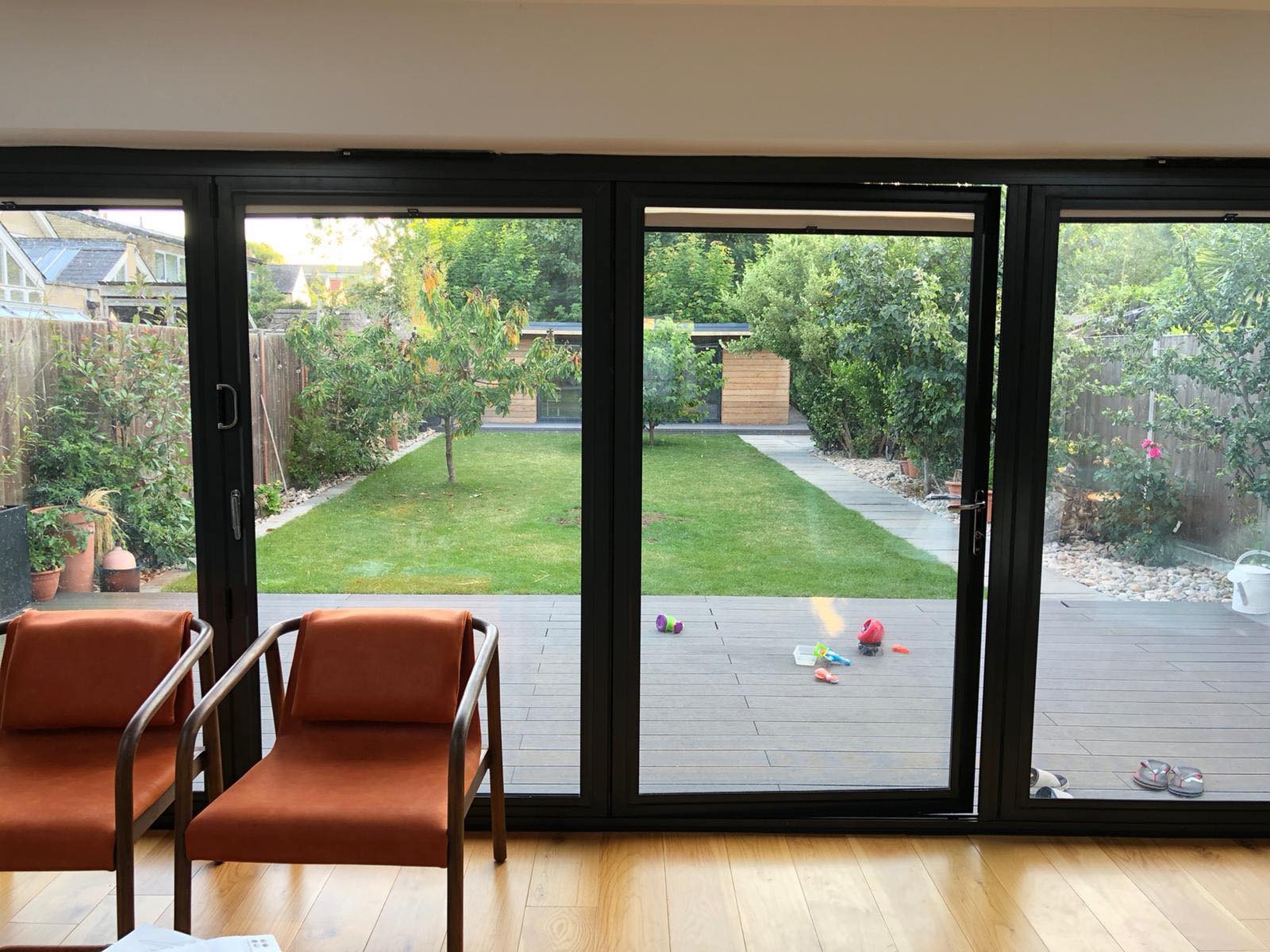 Widok na zadbany ogród z równo przystrzyżonym trawnikiem i drewnianym domkiem narzędziowym, widziany przez otwarte, nowoczesne, czarne, składane drzwi tarasowe z drewnianym tarasem i porozrzucanymi...