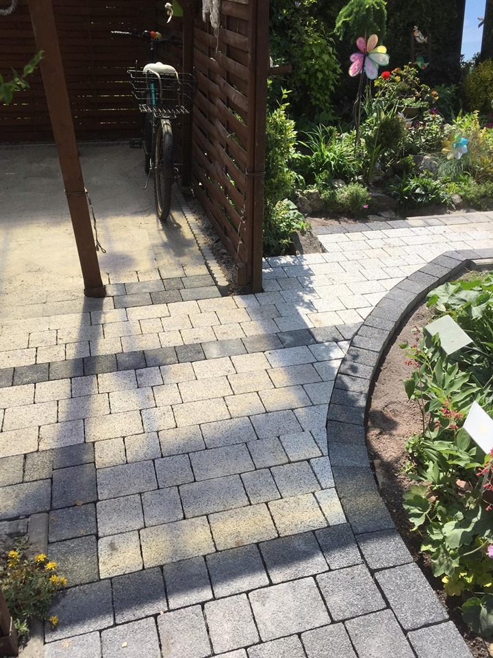 Ścieżka z kostki brukowej w odcieniach szarości i beżu prowadząca przez ogród z dekoracyjnymi wiatraczkami i rowerem opartym o drewniany płot.