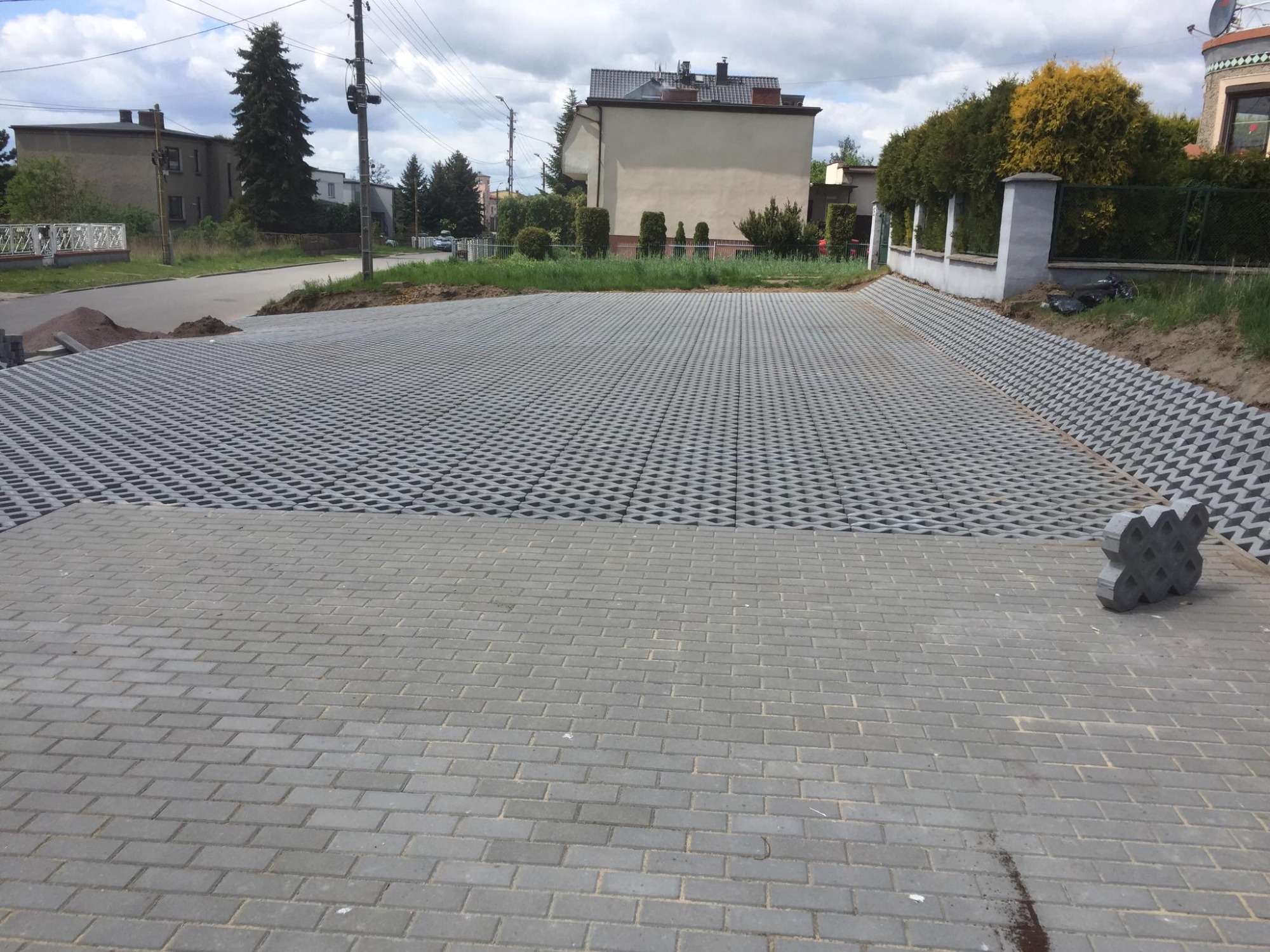 Fragment podjazdu wyłożonego dwoma rodzajami szarej kostki: standardową oraz ażurową typu 'plaster miodu', z widocznym stosem kostki ażurowej, przygotowany teren pod dalsze prace, w tle budynki...