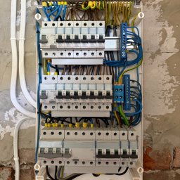 Inter-Style - Otwarta rozdzielnica elektryczna z rzędami bezpieczników Legrand i uporządkowanym okablowaniem w kolorowych izolacjach, zamontowana na częściowo otynkowanej ścianie z widoczną cegłą.
