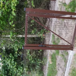 Otwarta, brązowa bramka z siatki metalowej na betonowej podmurówce, widoczna na tle bujnej zieleni.