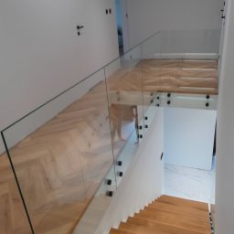 Zybi-pol zakład usługowo produkcyjny - Nowoczesne schody z drewnianymi stopniami i szklaną balustradą mocowaną na czarnych punktowych uchwytach, widok z góry na jasne wnętrze domu.