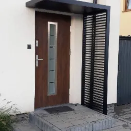 Nowoczesne zadaszenie nad wejściem do domu, z metalową konstrukcją w kolorze czarnym i drewnianymi drzwiami z elementami szkła. Schody wejściowe wyłożone szarą kostką.