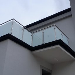 Zybi-pol zakład usługowo produkcyjny - Szklana balustrada z nierdzewnymi elementami na balkonie budynku o szarej elewacji, widok z dołu, fragment rusztowania z lewej strony kadru.