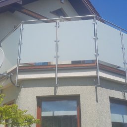 Zybi-pol zakład usługowo produkcyjny - Szklana balustrada balkonowa z nierdzewnymi słupkami na tle błękitnego nieba, widoczna antena satelitarna i fragment zieleni. Kąt ujęcia od dołu podkreśla nowoczesny design.