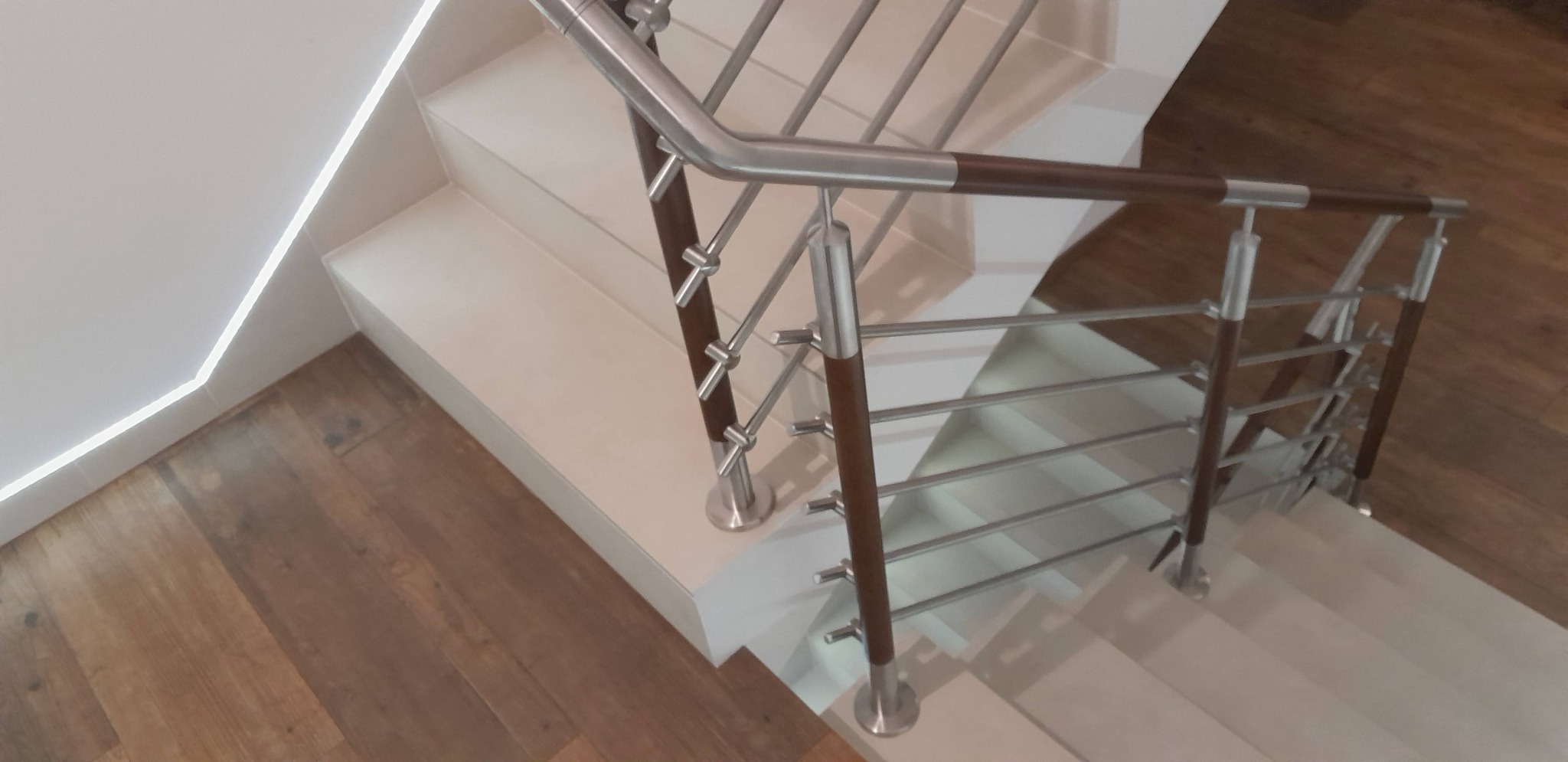 Widok z góry na schody z białymi stopniami i nowoczesną balustradą z elementami stali nierdzewnej i drewna w kolorze ciemnego brązu, widoczna podświetlana listwa przypodłogowa i parkiet.