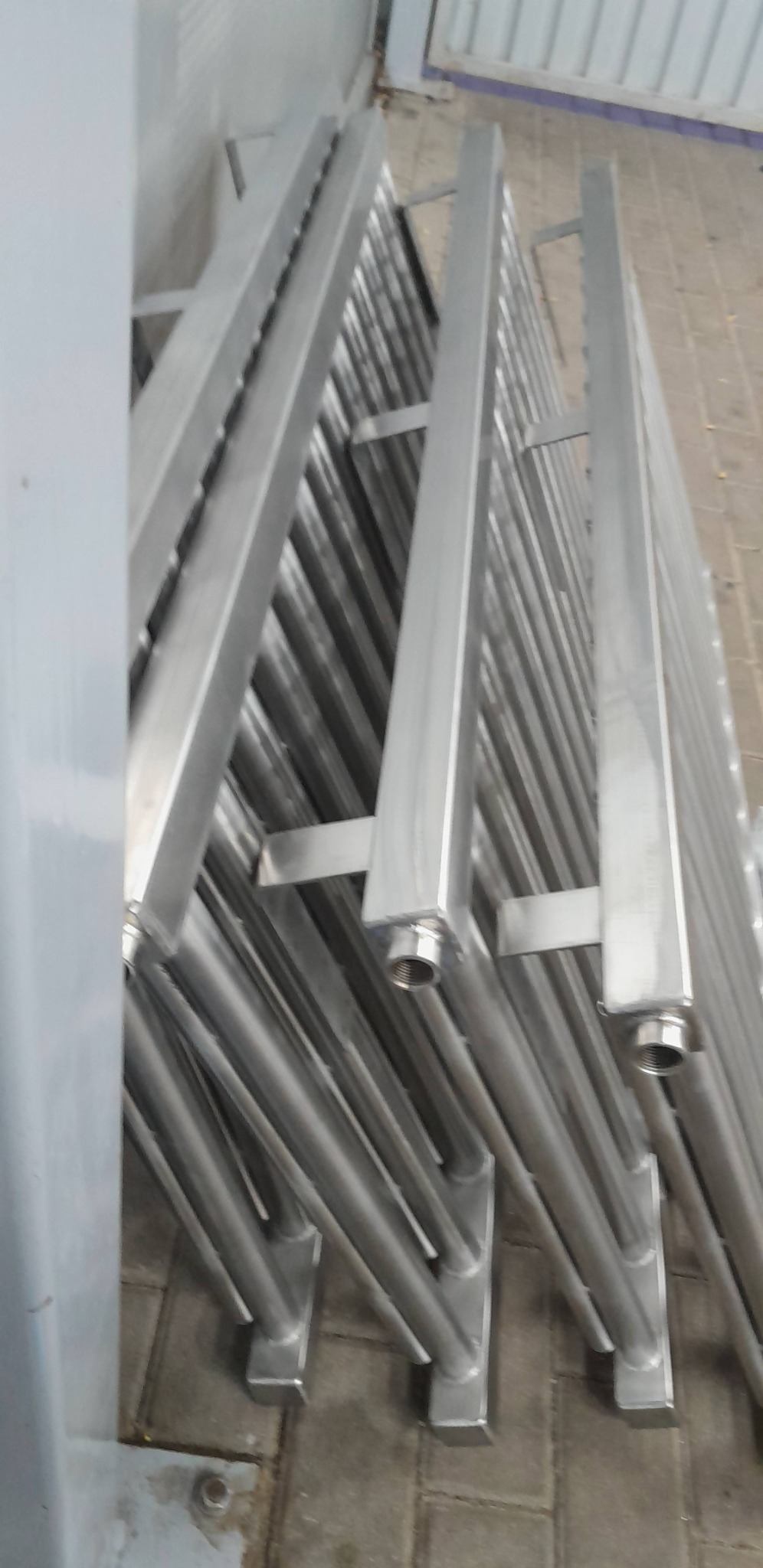 Stalowe, chromowane grzejniki drabinkowe ułożone jeden obok drugiego, widoczne gwintowane końcówki do podłączenia.