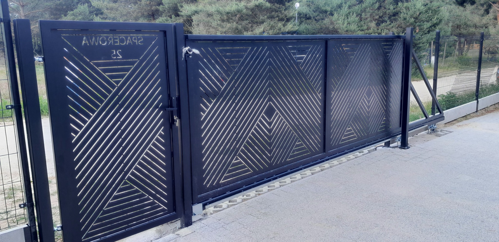 Czarna, przesuwna brama wjazdowa z geometrycznym wzorem szczelin, widoczny zamek i mechanizm przesuwny, na tle ogrodzenia z siatki i zieleni.
