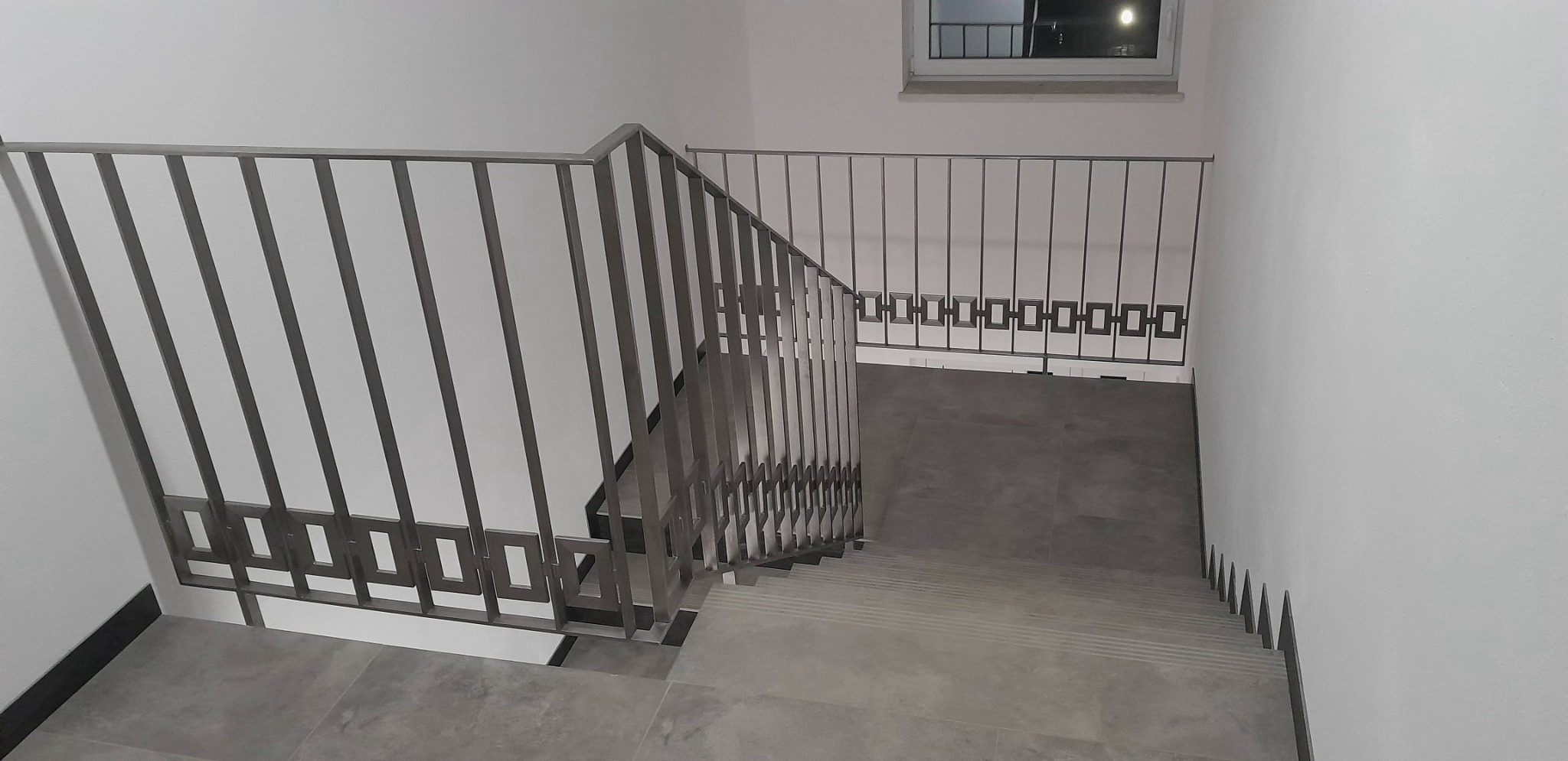 Widok z góry na schody z metalową balustradą o pionowych szczeblach i dekoracyjnych kwadratach, szare płytki na podłodze i schodach, białe ściany, okno w tle.