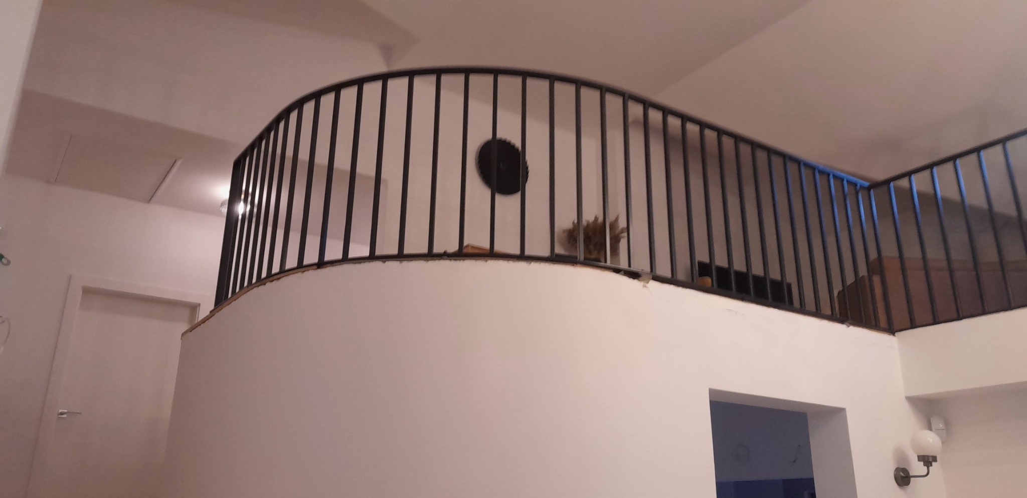 Wnętrze domu z czarną, metalową balustradą na antresoli o zaokrąglonym kształcie, widok z dołu, z elementami dekoracyjnymi na tle białych ścian.