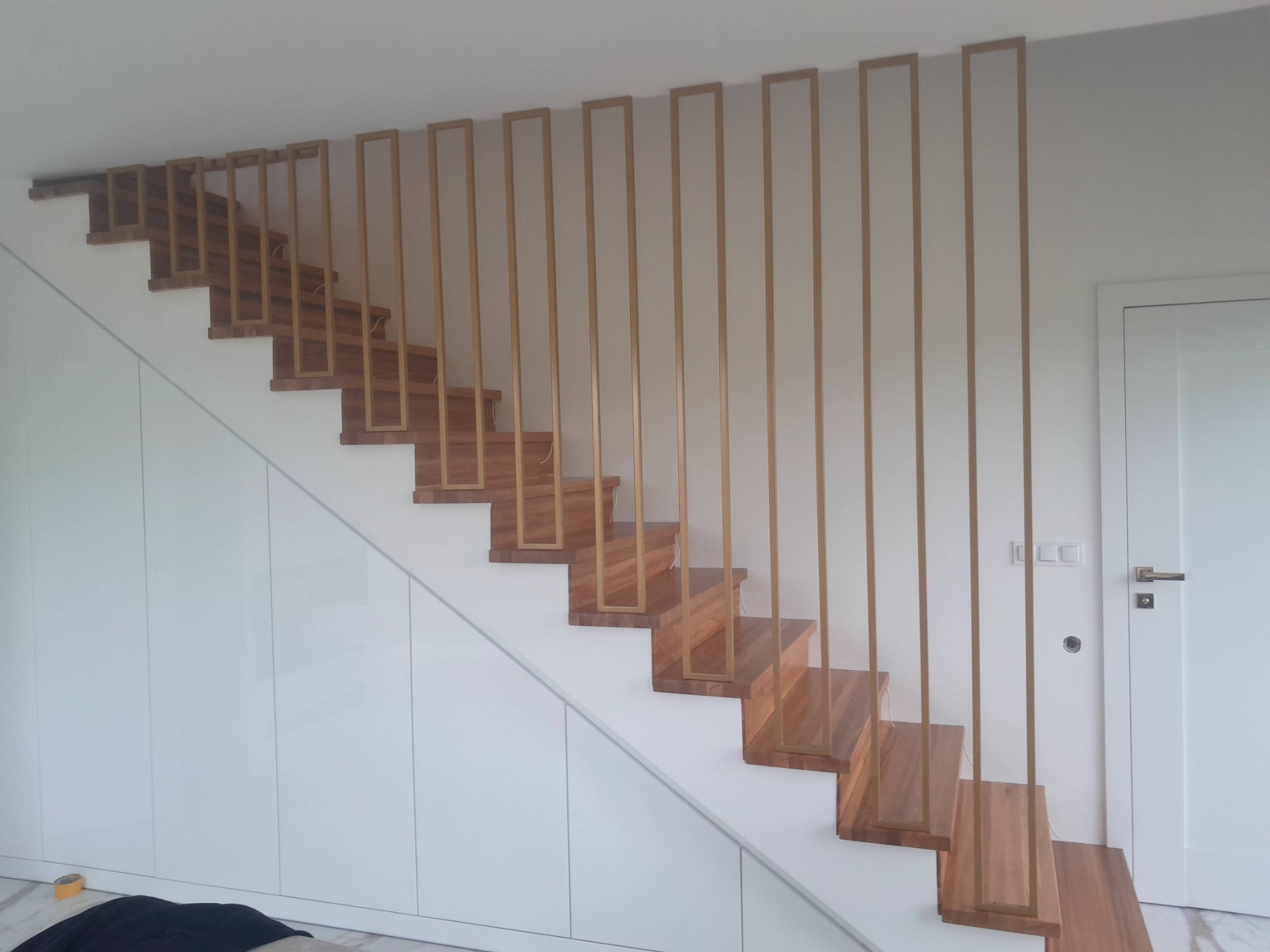 Nowoczesne schody drewniane z minimalistyczną, złotą balustradą w kształcie prostokątnych ramek, wkomponowane w białą zabudowę szaf wnękowych, oświetlone naturalnym światłem.