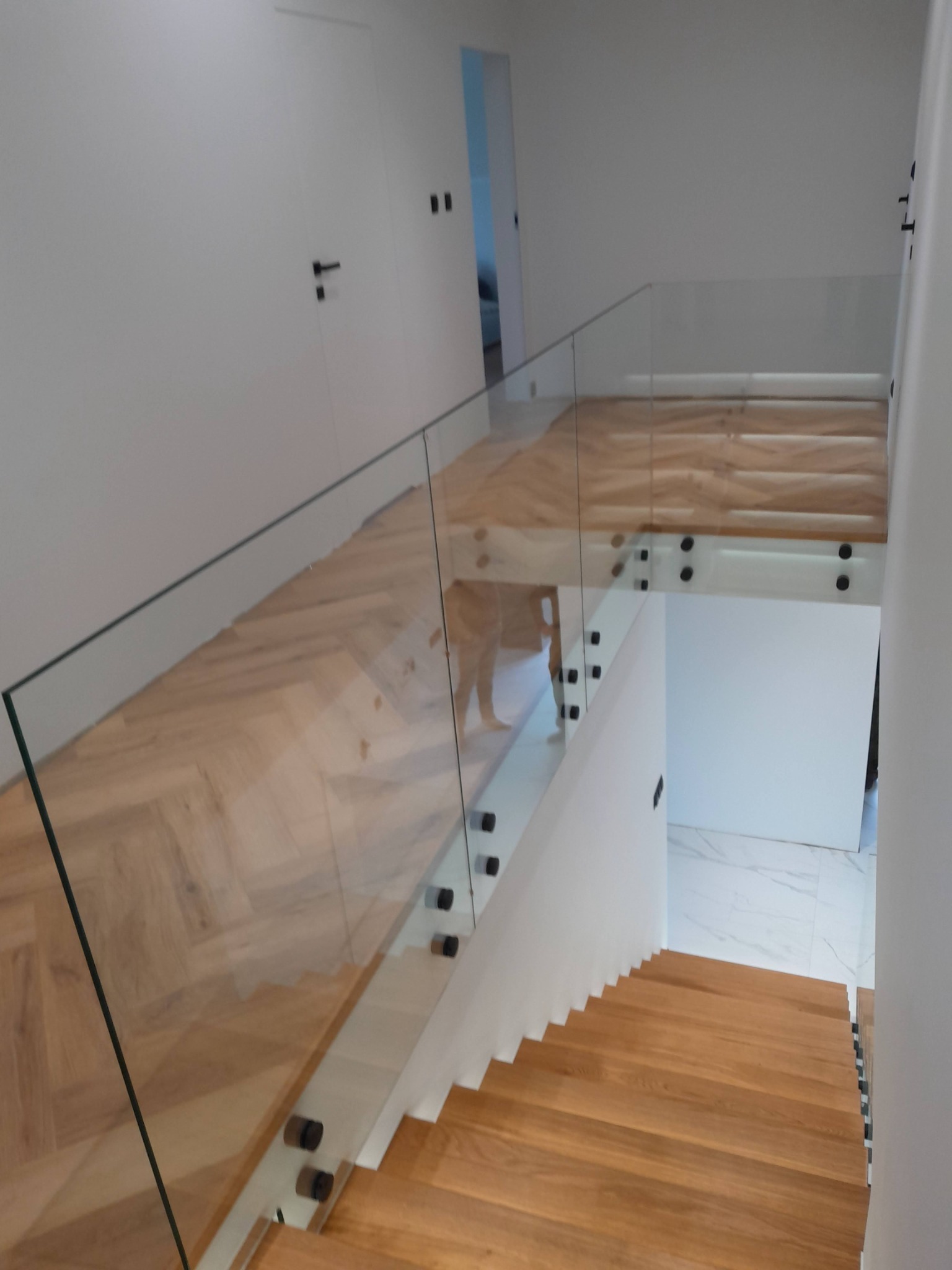Nowoczesne schody z drewnianymi stopniami i szklaną balustradą mocowaną na czarnych punktowych uchwytach, widok z góry na jasne wnętrze domu.