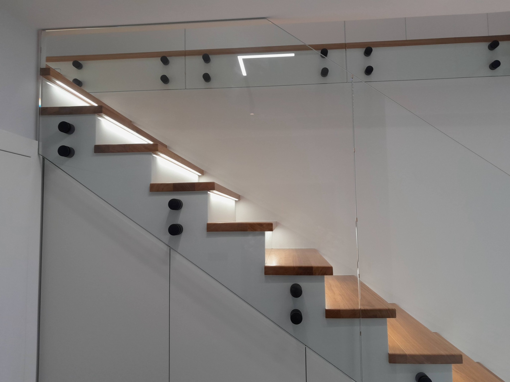 Nowoczesne schody z drewnianymi stopniami, podświetleniem LED i szklaną balustradą mocowaną na czarnych uchwytach punktowych.